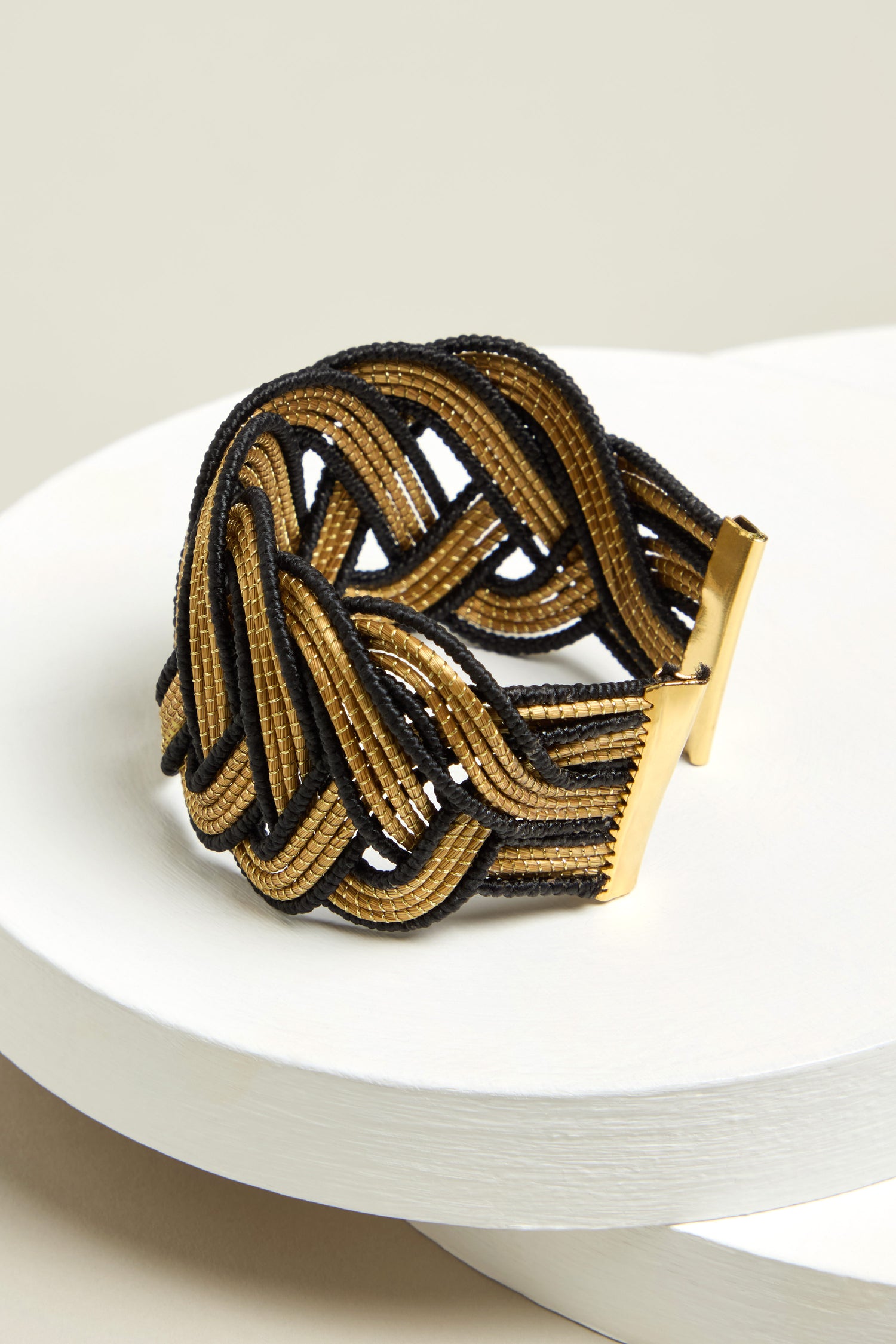 The Gold and Black Grass Woven Bracelet, showcasing a sustainable design, elegantly rests on a white round surface.