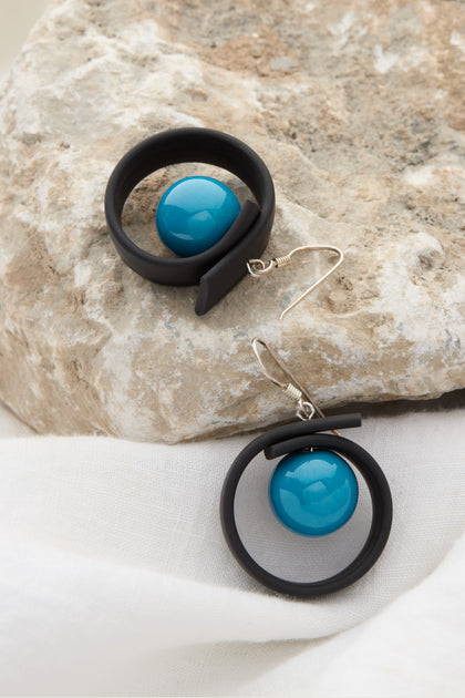 The Tempo Earrings, featuring black circular hoops and turquoise beads crafted from durable materials, are elegantly displayed against a textured rock and white fabric background.