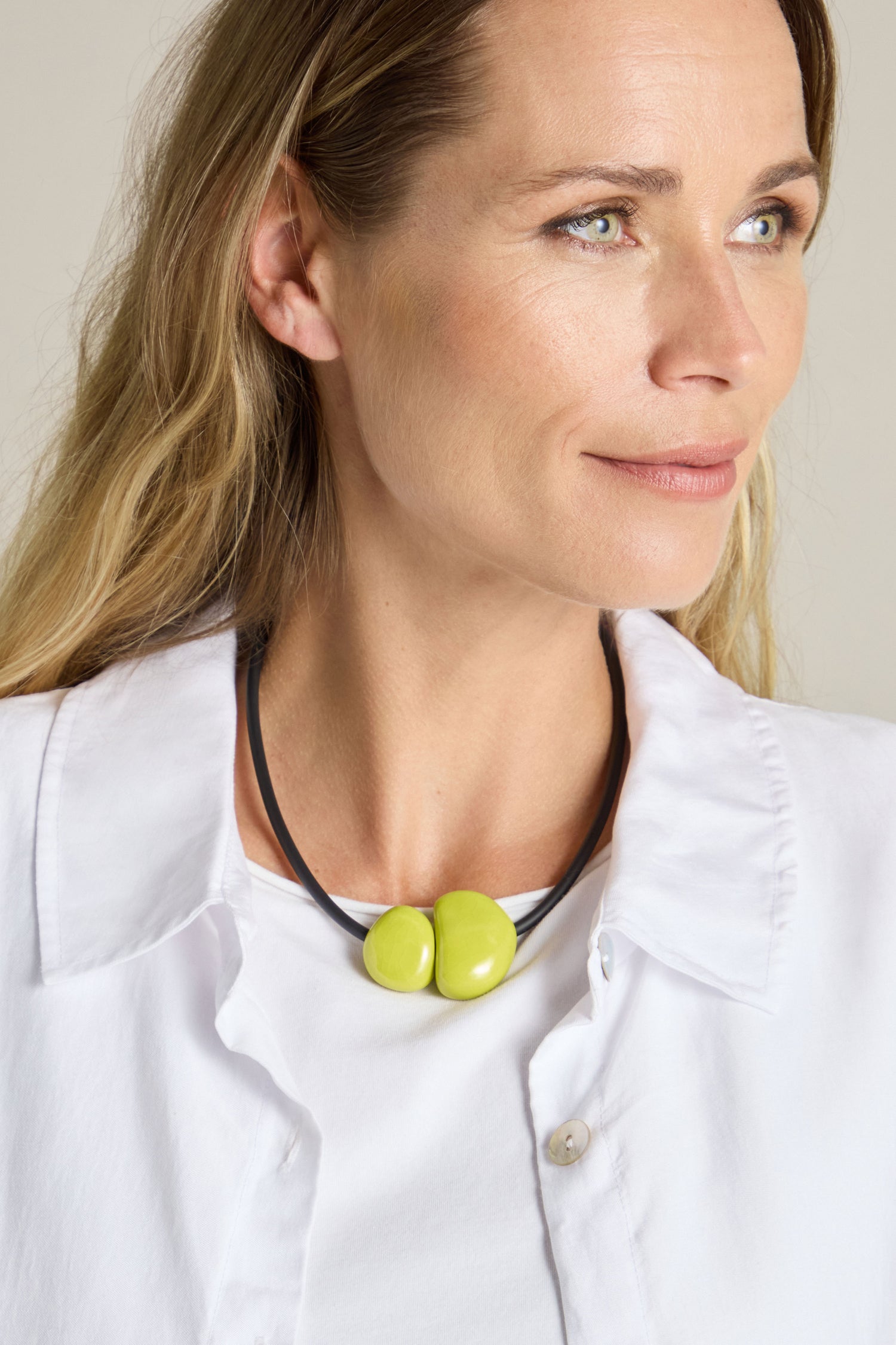 A person with long hair wearing a white shirt and a Short Ceramic Pebble Necklace featuring two large green beads and a magnetic clasp looks slightly to the side.