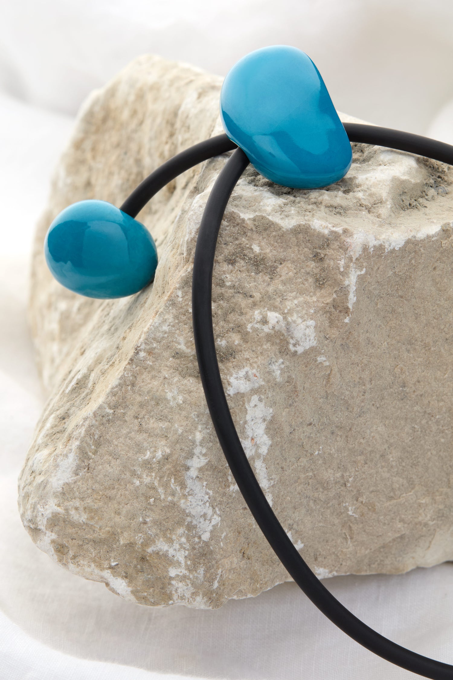 A Short Ceramic Pebble Necklace, featuring a black cord adorned with two turquoise stones, is elegantly displayed over a rough, light-colored rock, exuding contemporary style.