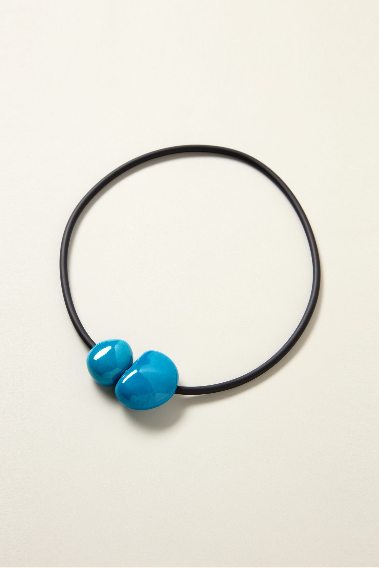 The Short Ceramic Pebble Necklace is a black circular ceramic piece adorned with two blue ceramic beads, set against a beige background. It includes a magnetic clasp for effortless wearing and boasts a contemporary design.