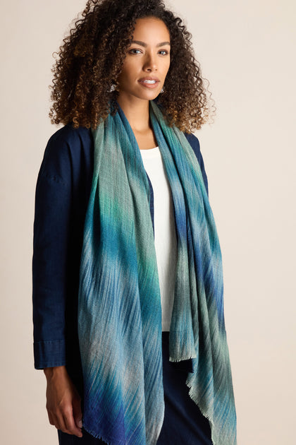 A woman with curly hair wearing a blue and green Sky Wool Cashmere Blend Scarf stands against a neutral background, her dark outfit contrasting nicely with the scarf’s herringbone stitch print.