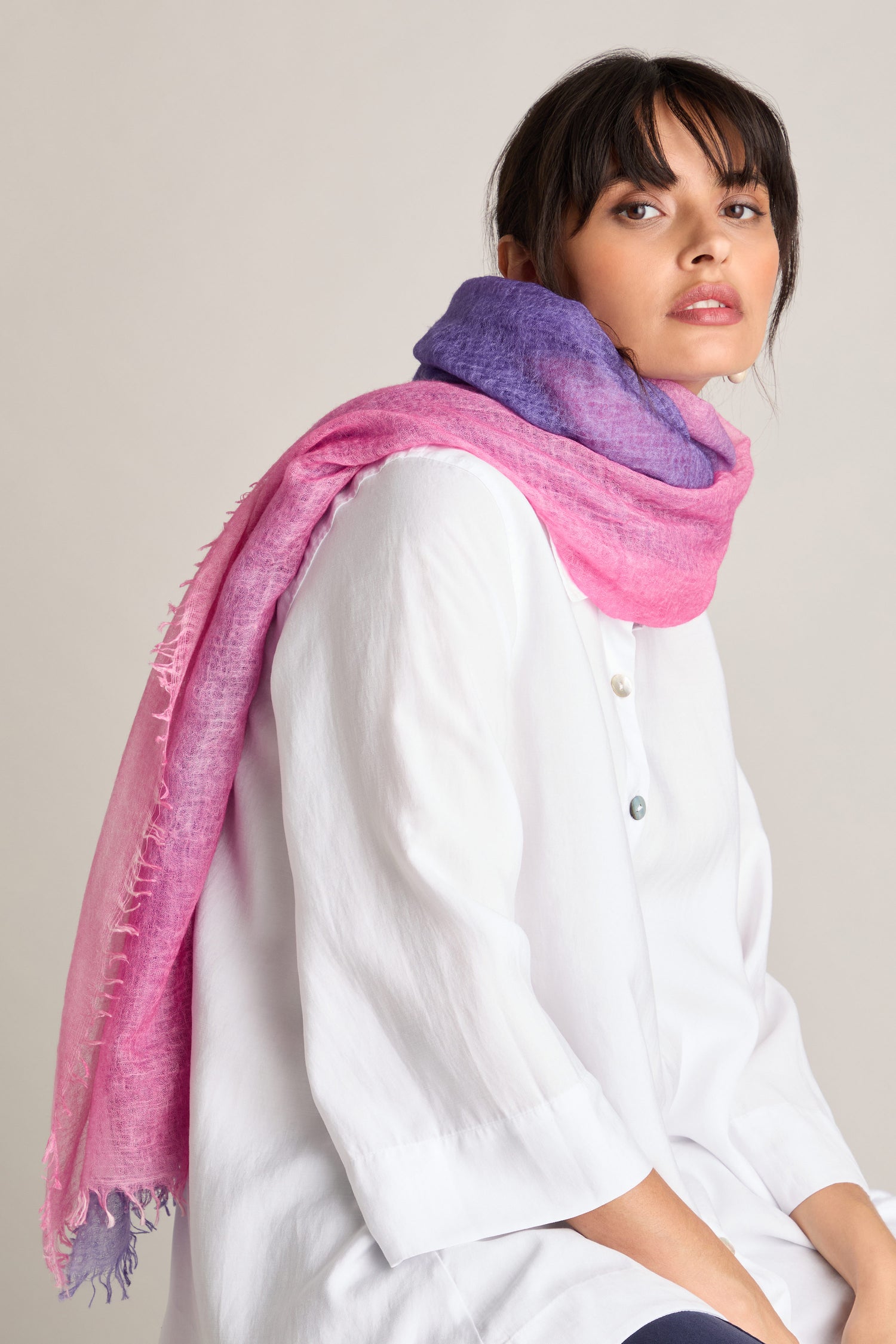 A person wearing a white shirt and the elegant Harmony Cashmere Scarf, an autumn accessory in pink lilac, sits against a beige background.