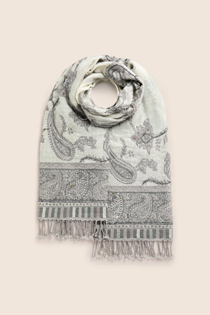 The Paisley Sequin Wool Scarf is a light gray, neutral-toned accessory made of 100% wool, adorned with paisley and floral motifs and finished with fringed edges. It is shown folded in a circular arrangement on a beige background.