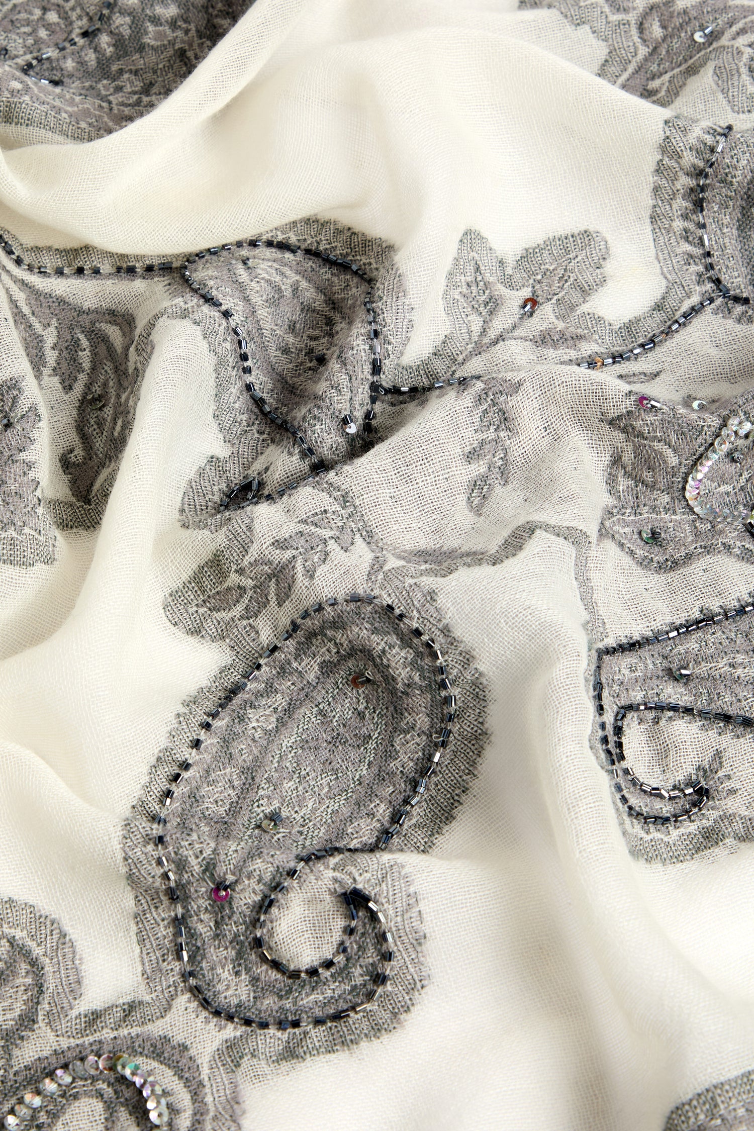 A close-up image of the Paisley Sequin Wool Scarf. The fabric is light-colored with intricate gray paisley motifs and floral patterns, adorned with small beads.