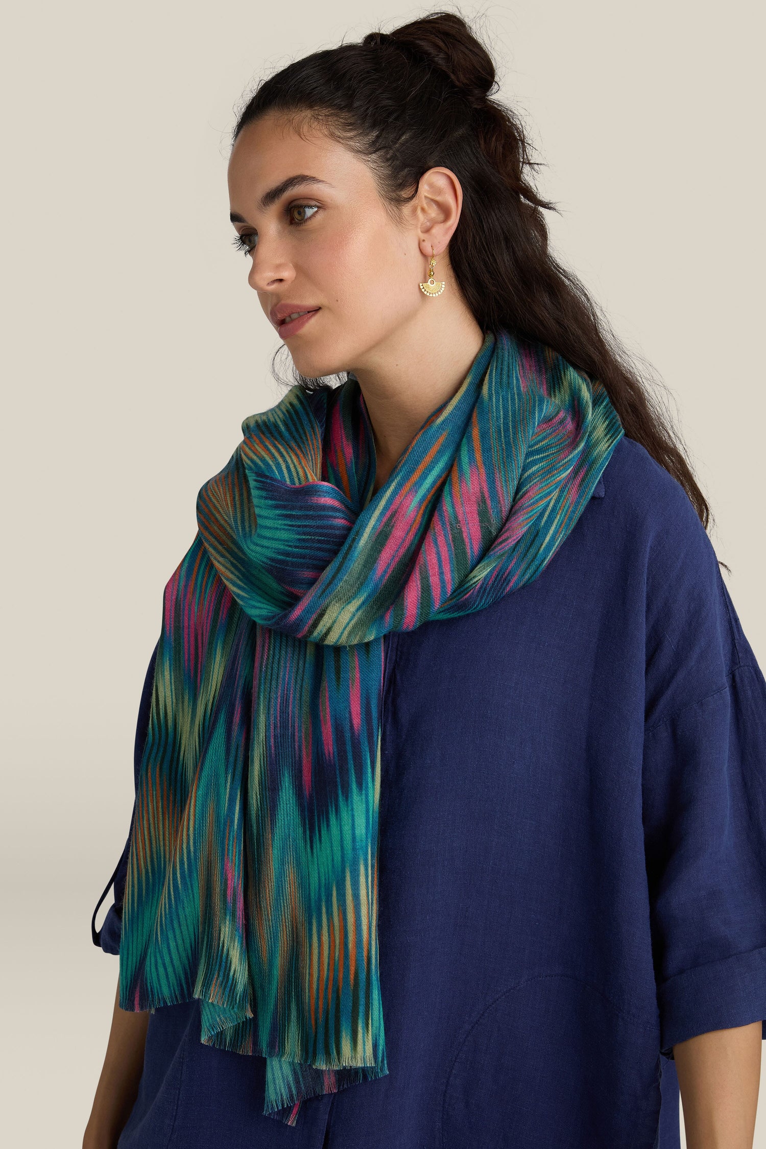 A woman wearing a blue top and an Autumnal Ikat Wool Scarf featuring colorful, ikat-inspired patterns gazes to the side against a neutral background.