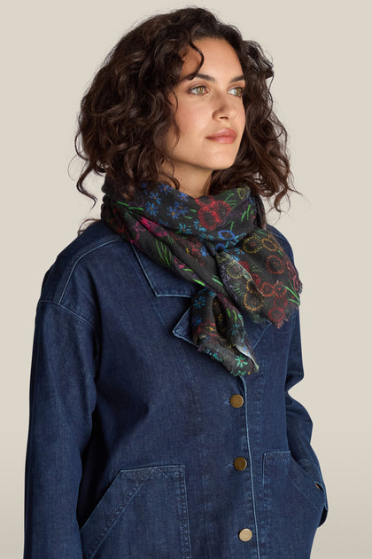 A person with curly hair, dressed in a denim jacket and a Winter Floral Wool Scarf featuring an embroidered floral print, gazes to the side against a plain background.