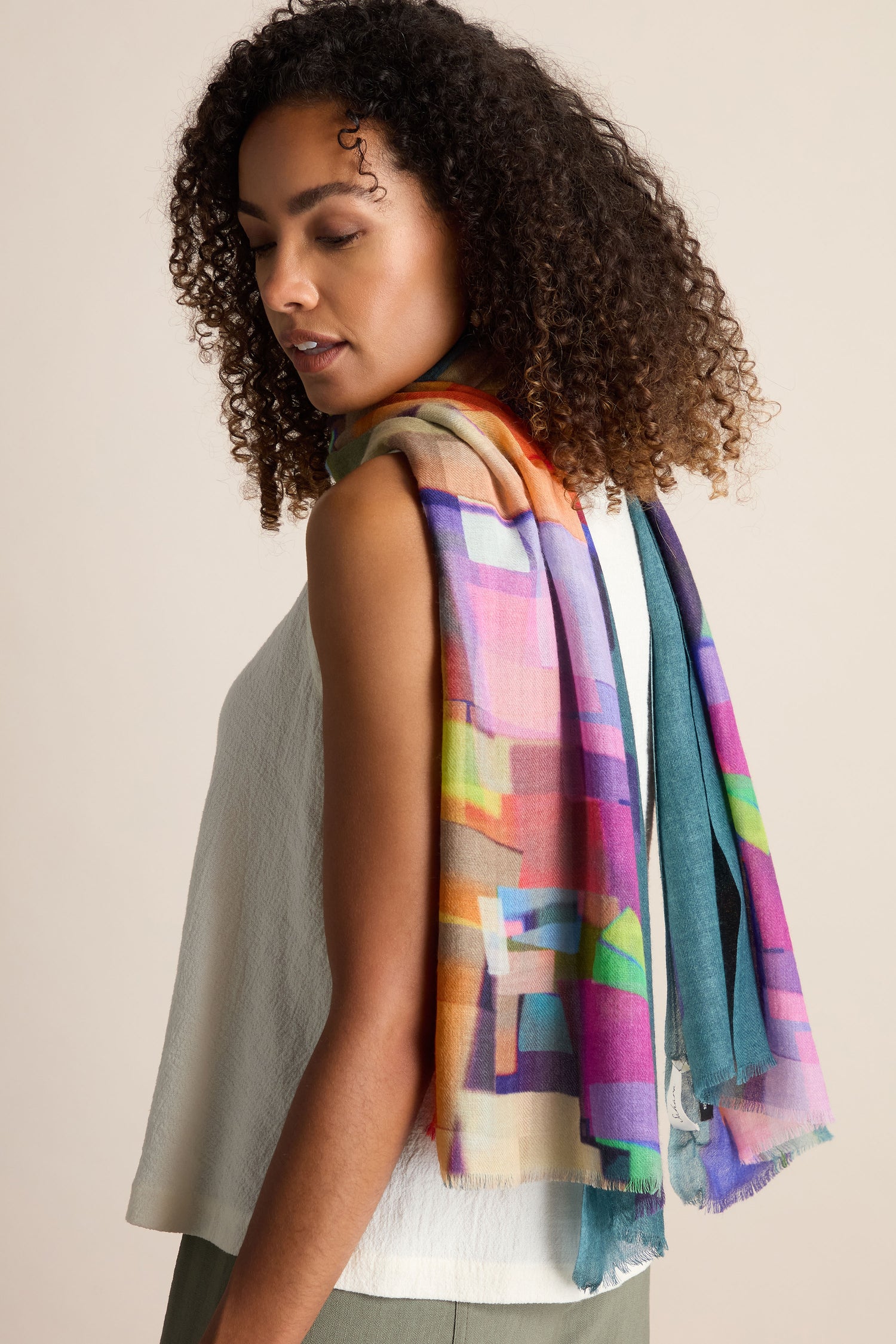 A woman with curly hair wearing a sleeveless top and a vibrant Square Collage Wool Scarf draped over her shoulders, looking down to her left.