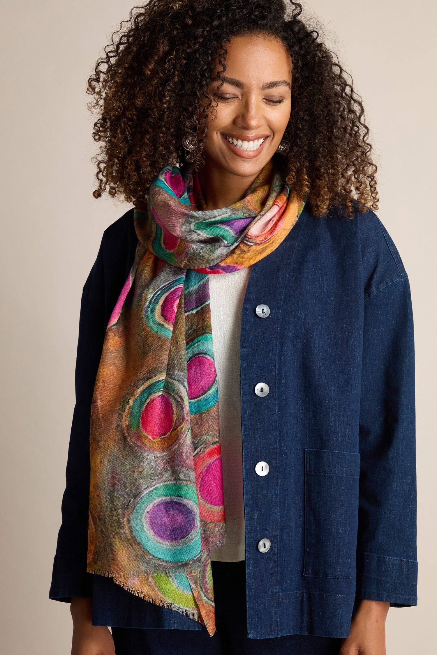 A person with curly hair smiles while wearing a vibrant Watercolour Circles Wool Mix Scarf and a blue buttoned jacket over a white top.