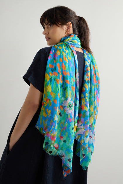 Wearing an Abstract Painted Garden Scarf over a dark dress, someone glances over their shoulder against a plain backdrop.