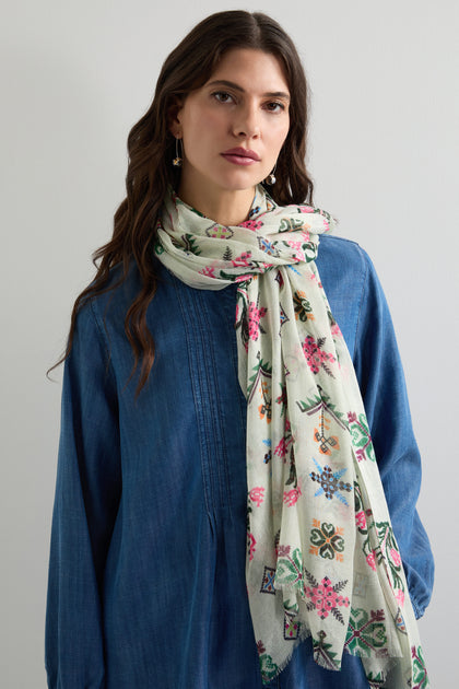 A person in a blue denim dress wears a Cross Stitch Print Scarf with colorful floral patterns, its traditional embroidery adding heritage charm against a plain background.