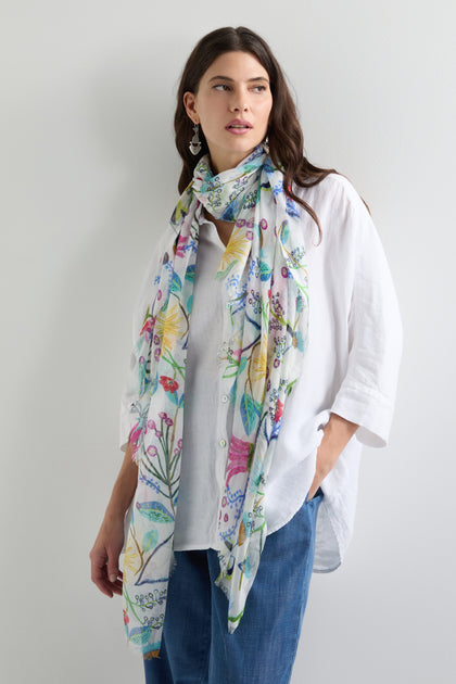 Against a plain background, a person wears a white shirt, blue jeans, and the vibrant Wild Garden Scarf adorned with a floral design.