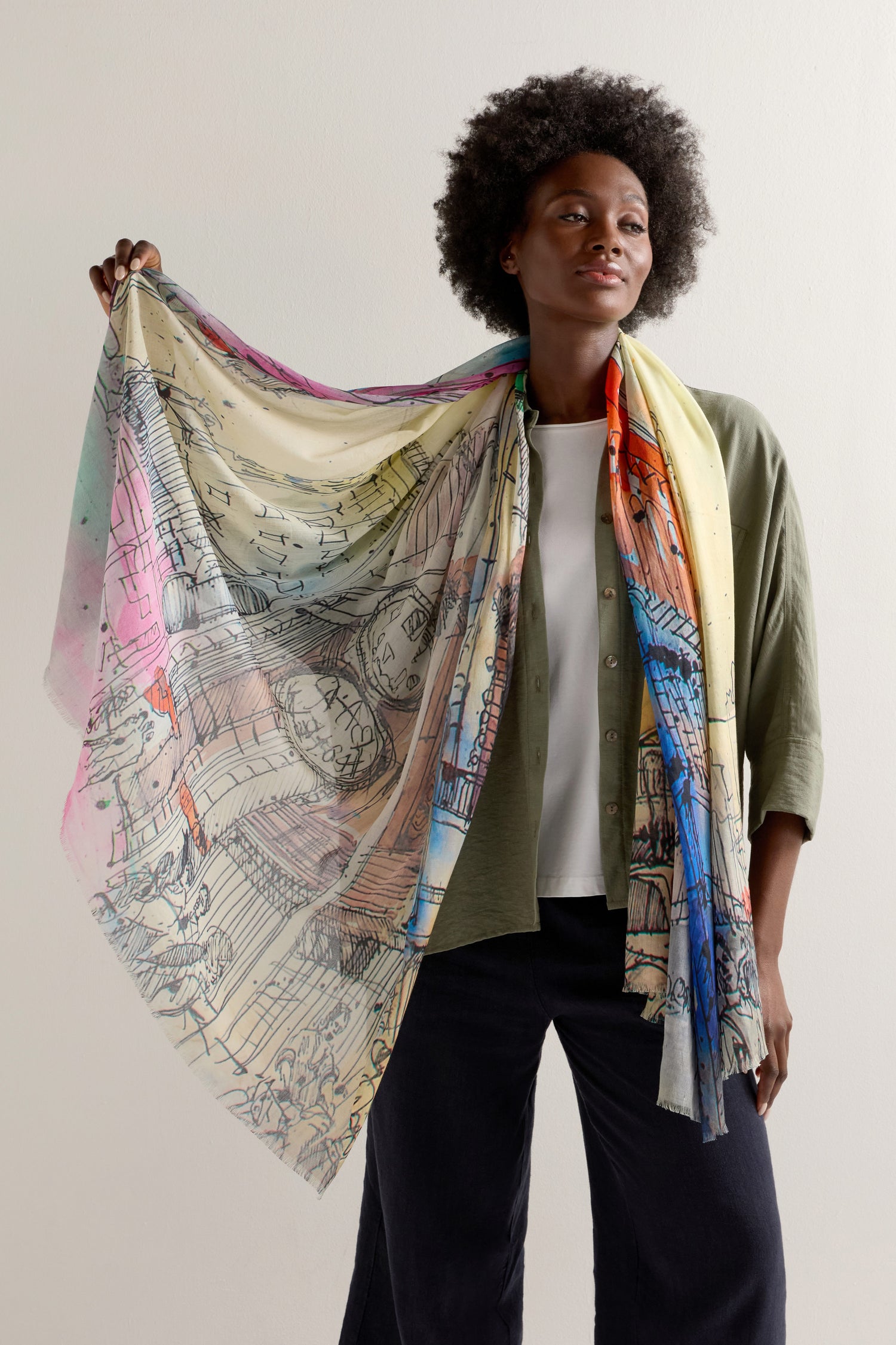 A person drapes an Abstract City Sketch Cotton Modal Scarf over their shoulder, vibrant and lively. They wear a green shirt with dark pants, adding an urban flair that complements the scarf's dynamic design.