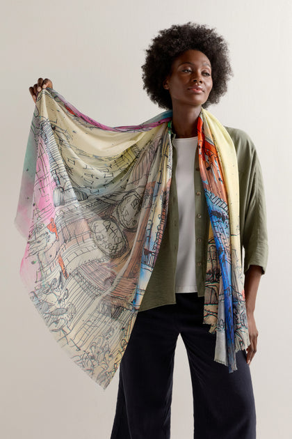 A person drapes an Abstract City Sketch Cotton Modal Scarf over their shoulder, vibrant and lively. They wear a green shirt with dark pants, adding an urban flair that complements the scarf's dynamic design.