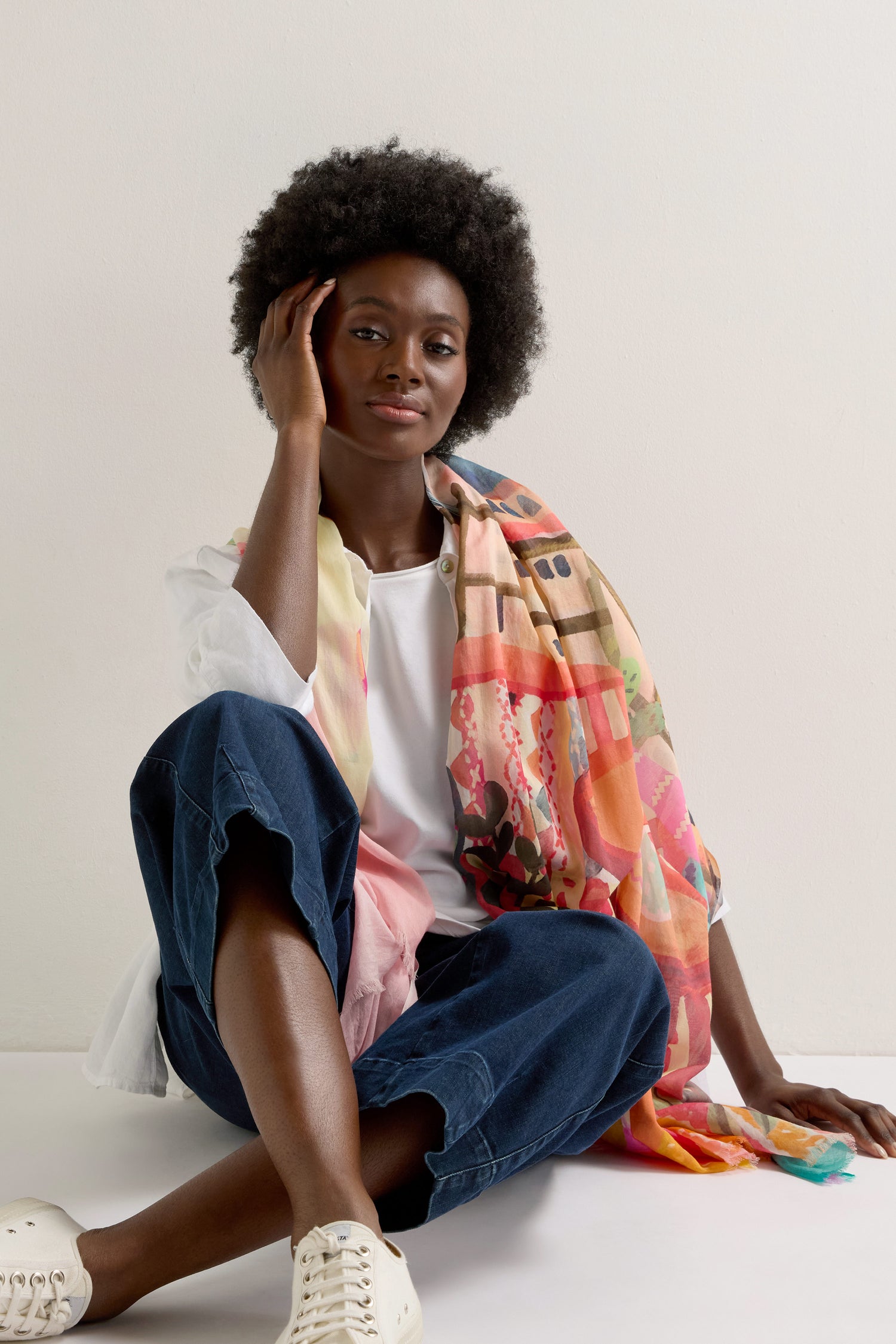 A person sits relaxed against a white wall, dressed in a white shirt and blue pants, accessorized with the Marrakech Cotton Modal Scarf. One hand touches their head, finishing the ensemble with stylish white sneakers.