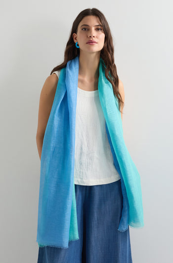 Against a light background, a person wears a white top, blue pants, and blue earrings. Their look is elevated with an Ombre Linen Mix Scarf in blue and teal shades, adding a stylish gradient effect.