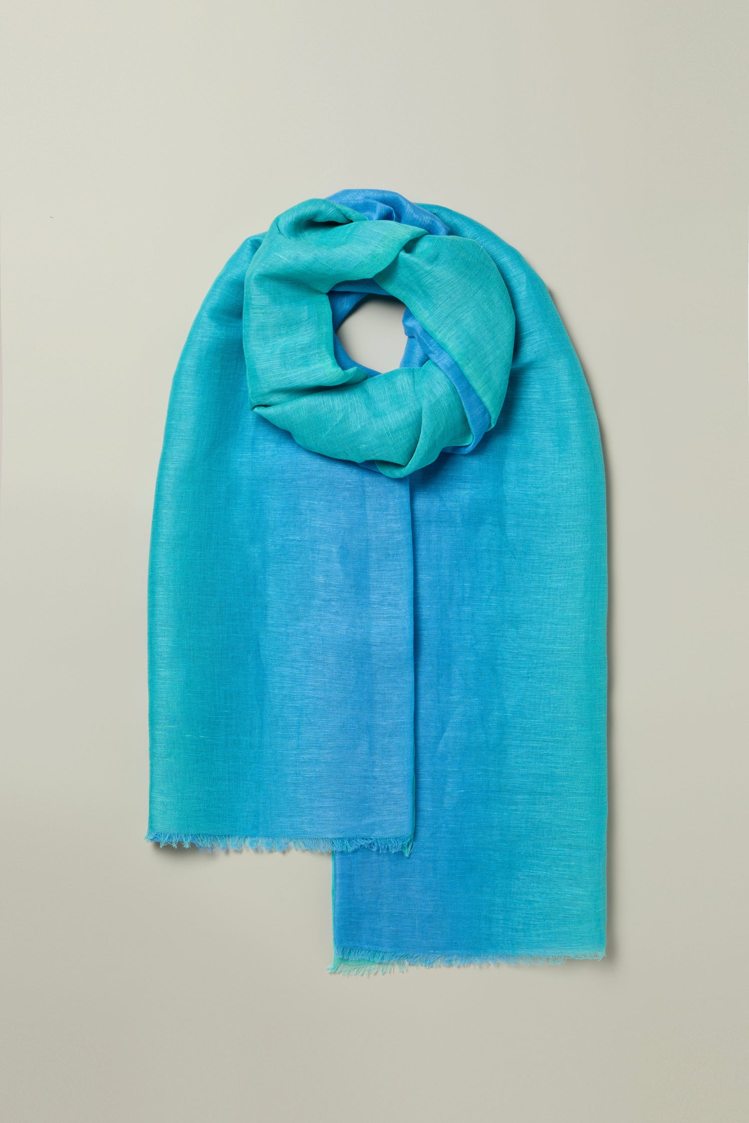 The Ombre Linen Mix Scarf, featuring a light gradient of blue and teal shades, is elegantly displayed on a soft grey backdrop.