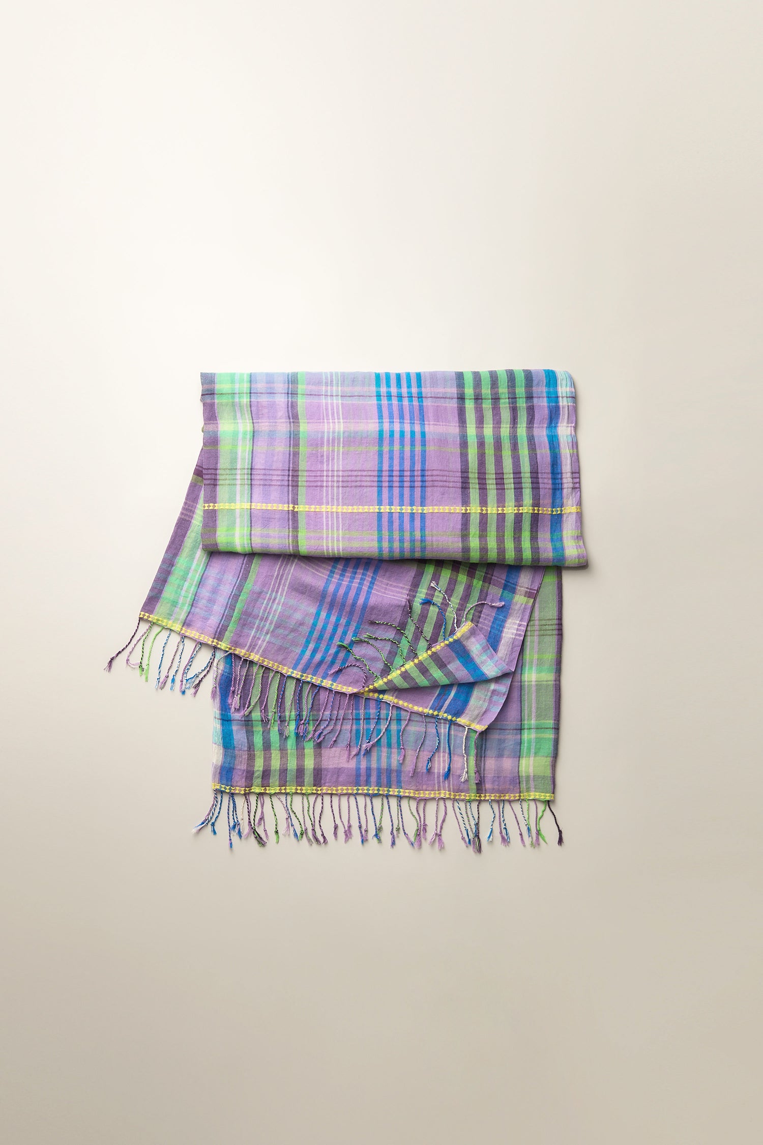 The Check Stitch Cotton Scarf, showcasing a vibrant check pattern in purple, blue, and green hues with fringed ends, elegantly displayed against a neutral backdrop.