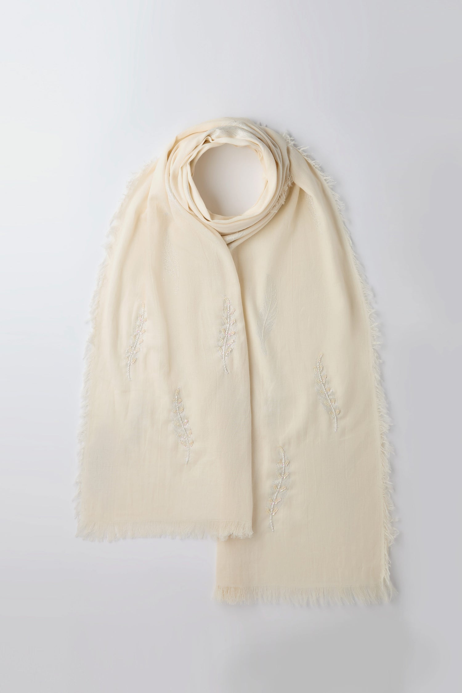 The Hand Embroidered Feather Scarf is cream-colored, featuring fringed edges and delicate hand embroidery patterns, crafted from a lightweight fabric.