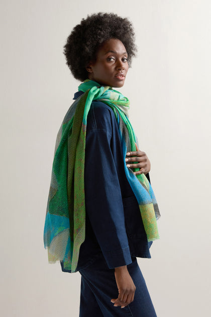 A person with an afro hairstyle wears a dark blue outfit and the Tide Sparkle Scarf, a lightweight green and blue accessory, enhancing their shimmering elegance against a plain light background.