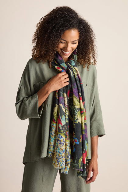 A person with curly hair is smiling and wearing a green outfit, accessorized with the Painted Garden Wool Mix Scarf adorned in a colorful botanical print. The background is plain and light-colored.