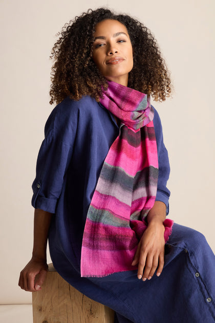 A woman with curly hair wearing a blue dress and a Crimson Twilight Stripe Scarf, glowing in rich sunset shades, sits on a wooden block against a neutral background. This ensemble is perfect for autumn and winter fashion.