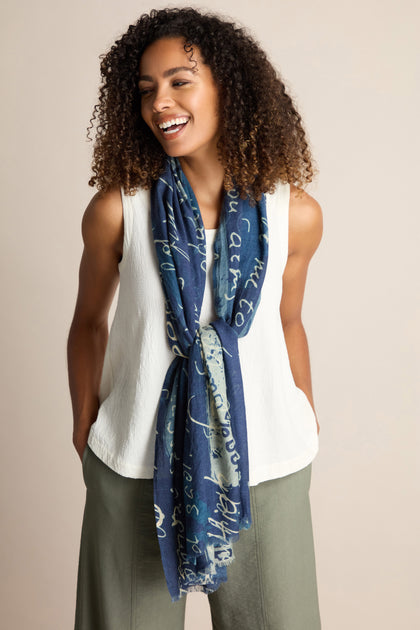 A person with curly hair wearing a white sleeveless top, green pants, and a long blue Love Language Printed Scarf stands against a plain background, smiling with hands in pockets.