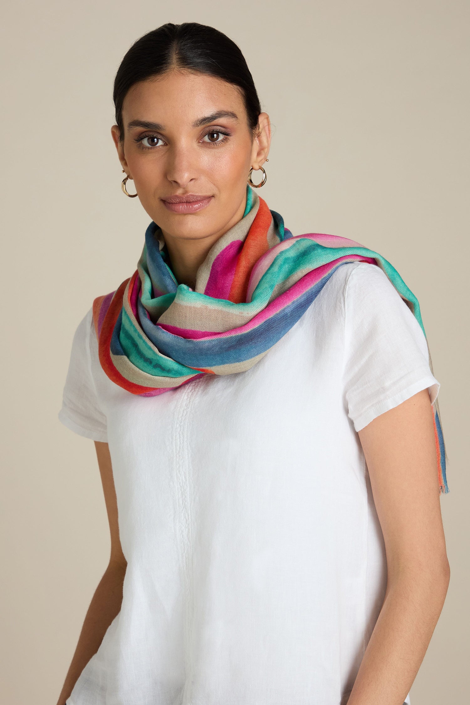 A woman with dark hair wears a white top and a Painterly Stripe Wool Mix Scarf adorned with colorful, brushstroke-inspired stripes.