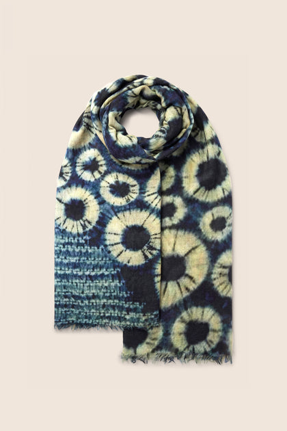 The Tie Dye Wool Scarf, showcasing a navy and cream floral pattern and artfully rolled design, features fringed ends and a soft texture. This versatile accessory is perfect for all-occasion styling.