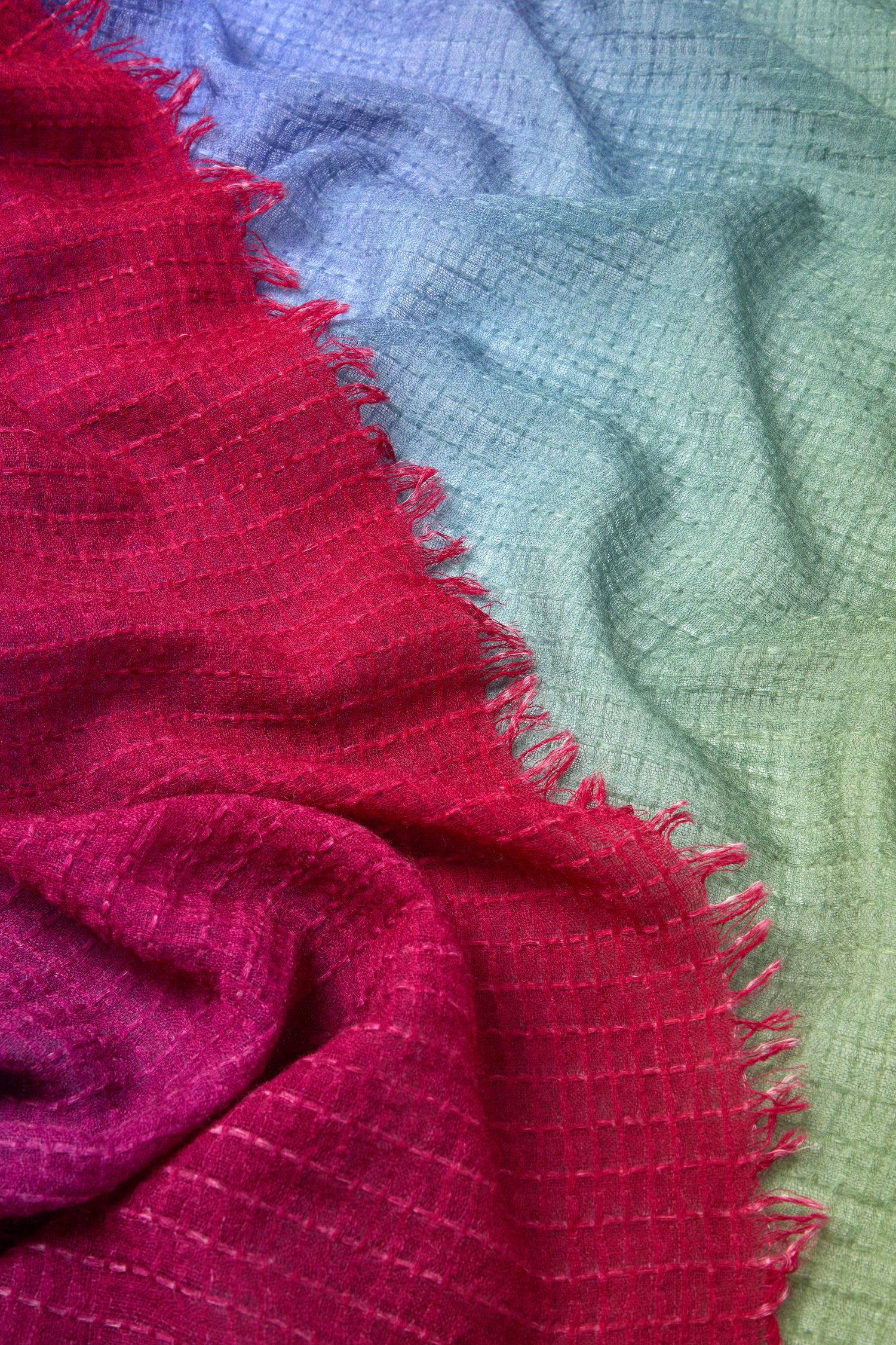 Close-up of the Soft Colour Blend Wool Scarf, showing its textured fabric with a gradient from red on the left to green on the right. The scarf features fringed edges and appears to be loosely woven.