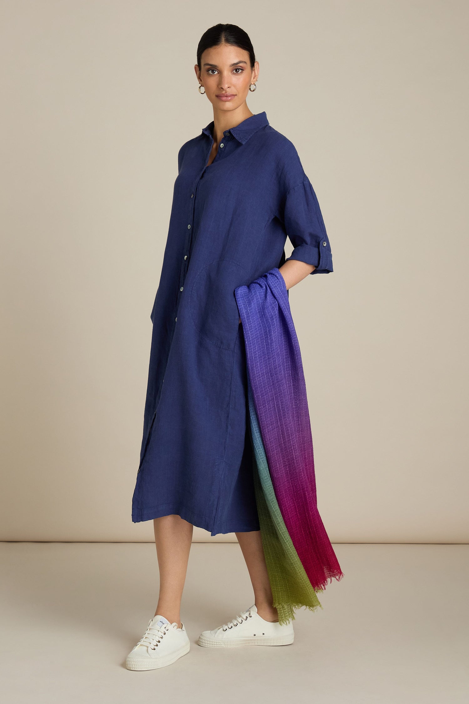 A person stands wearing a long-sleeve blue button-down dress paired with white sneakers. They hold the Soft Colour Blend Wool Scarf, which features an ombre of jewel tones from purple to green. With their hair tied back and standing against a beige background, they epitomize versatile, all-occasion styling.