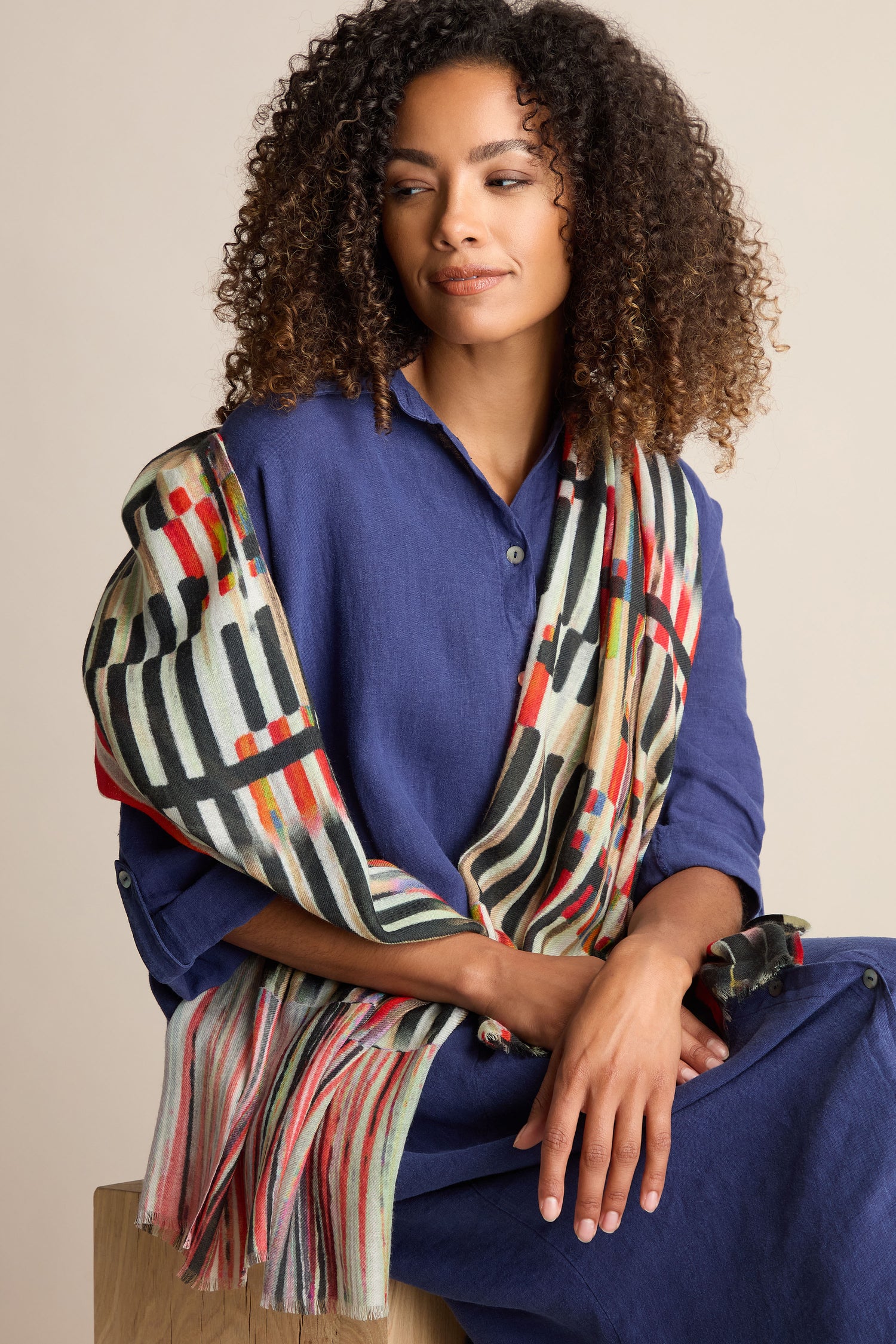 A woman with curly hair sits on a wooden stool, wearing a blue dress and the luxurious Pianist Wool Mix Scarf, adorned with vibrant geometric patterns.