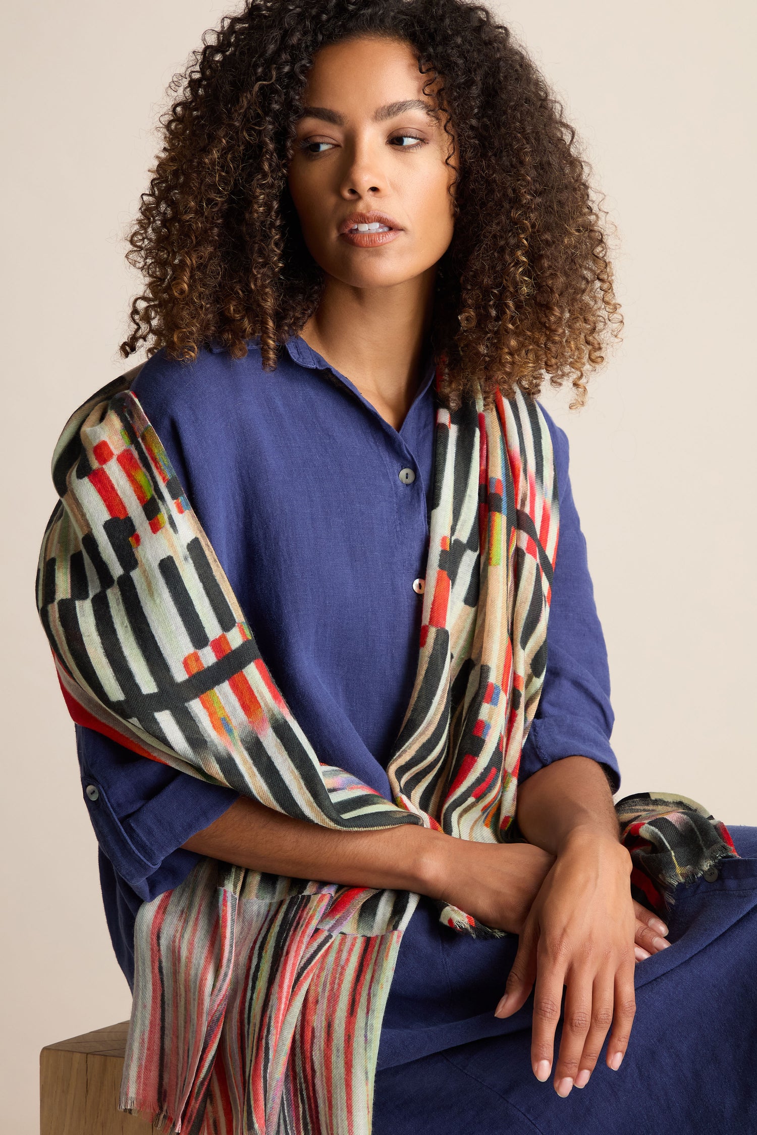 A person with curly hair wearing a blue button-up shirt and a Pianist Wool Mix Scarf, featuring a colorful pattern and piano key print, draped over their shoulder sits against a plain background.