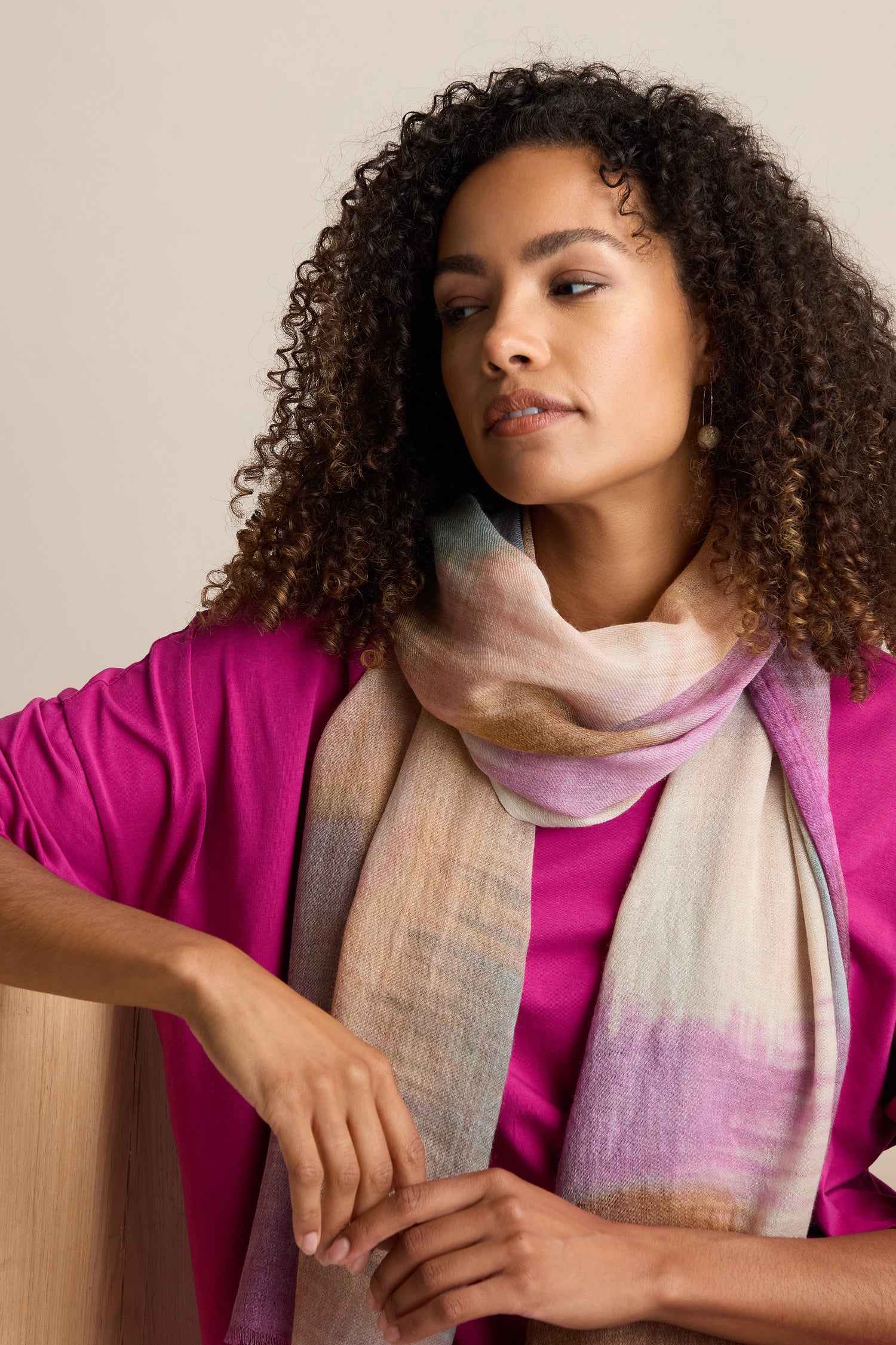 A person with curly hair, dressed in a pink top and the Colour Blend Circle Print Scarf, sits against a neutral background, glancing to the side. The scarf showcases a captivating watercolour-inspired design that brings an artistic flair to their outfit.