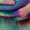 Close-up of a person's hands weaving the Colour Fusion Wool Mix Scarf, showcasing its colorful, abstract design. The scarf boasts vibrant hues including green, blue, purple, and pink, reminiscent of a watercolor print.