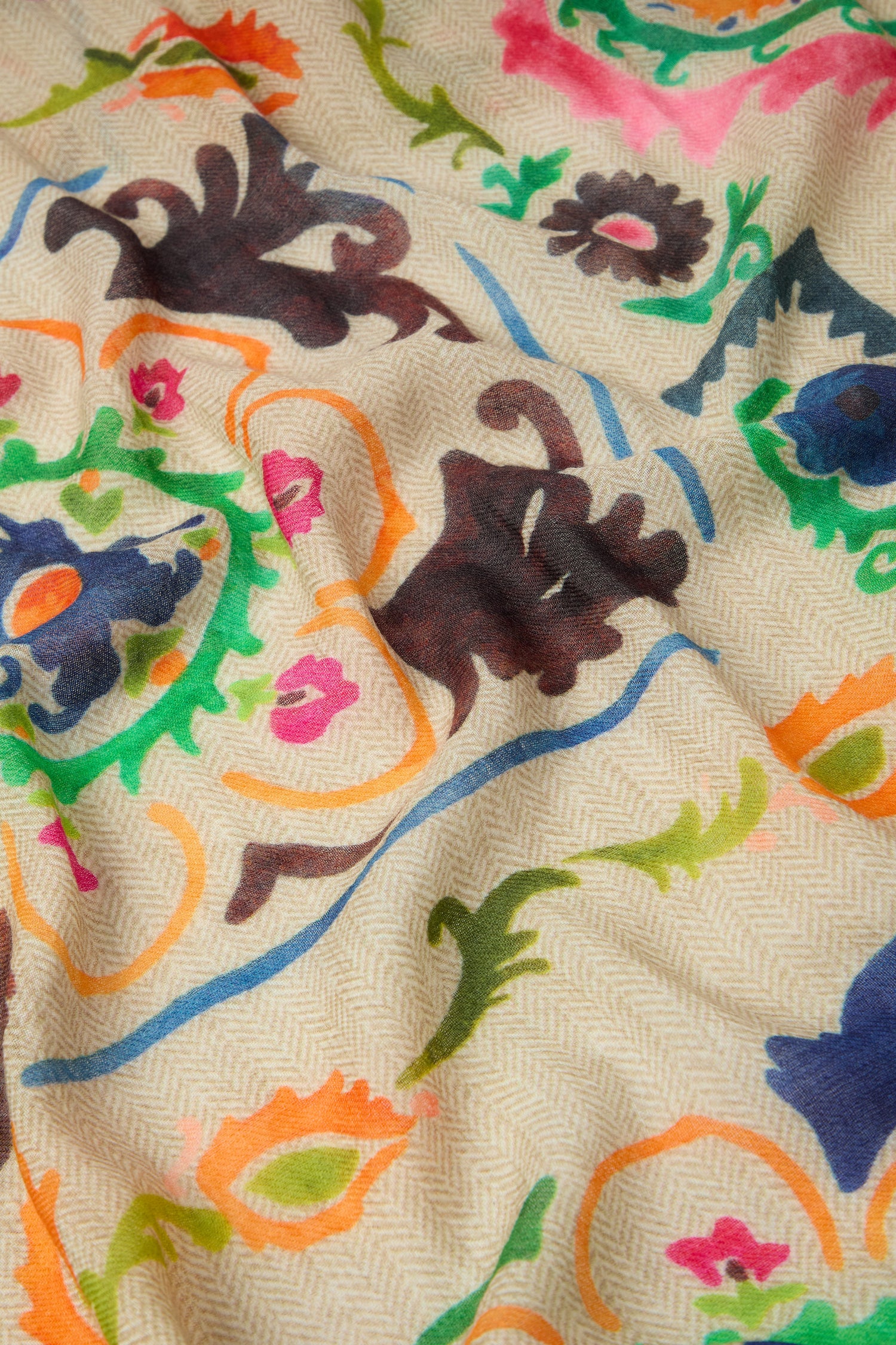 A close-up of the Painted Paisley Wool Mix Scarf, showcasing its light beige fabric with a herringbone pattern and a vibrant, multicolored floral and abstract design in shades of pink, green, blue, orange, and brown reminiscent of a hand-painted paisley scarf.