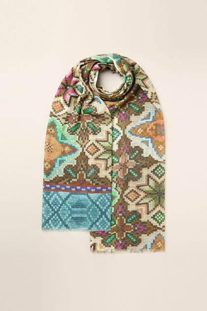 The Mosaic Print Wool Mix Scarf features an array of multicolored geometric and floral patterns, loosely rolled on a beige background.