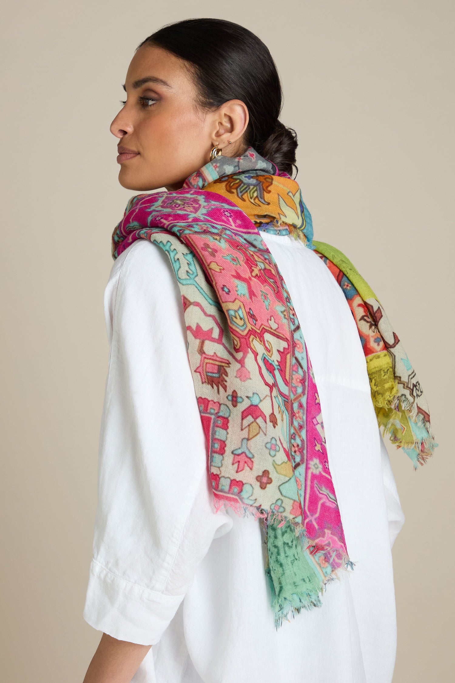 A woman in a white shirt adorned with the Medina Wool Scarf, featuring a vibrant Moroccan motif, draped over her shoulders, looks to the left against a plain backdrop.