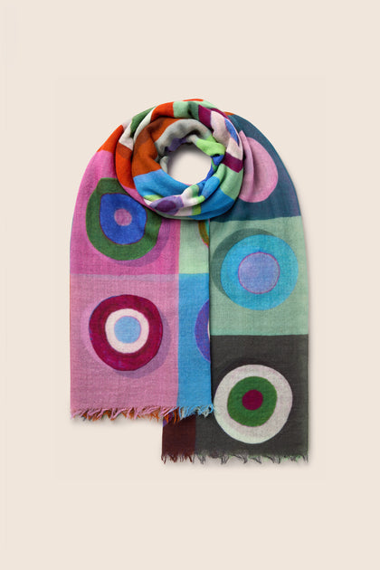 The Concentric Circles Wool Scarf features a multicolored design with bold concentric circles in shades of pink, blue, green, and orange set against a plain background. This geometric print accessory injects vibrant energy into any outfit.