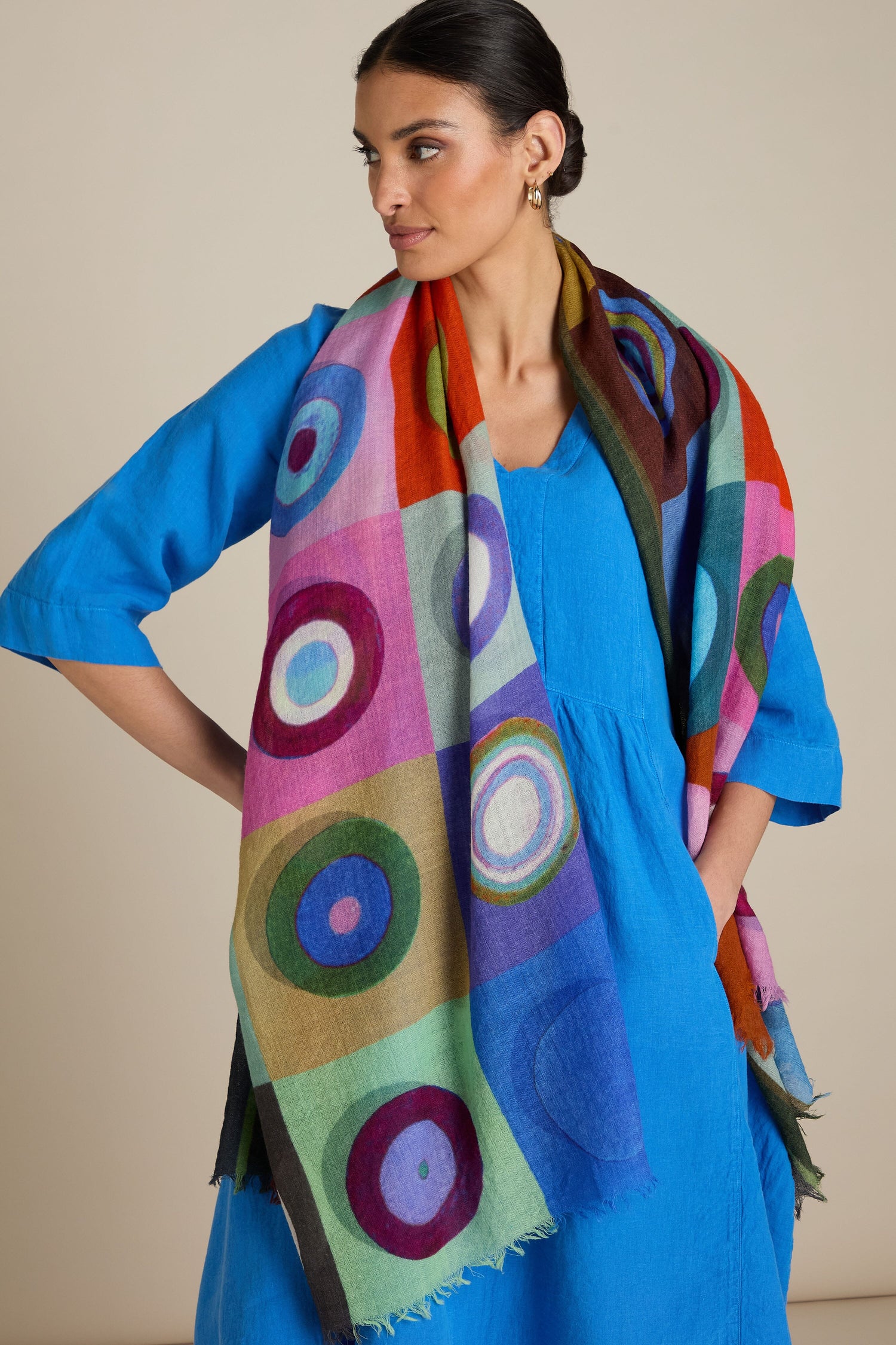 A woman in a blue dress poses with the Concentric Circles Wool Scarf, featuring a vibrant geometric print, draped over her shoulders, adding an eye-catching accessory to her ensemble.