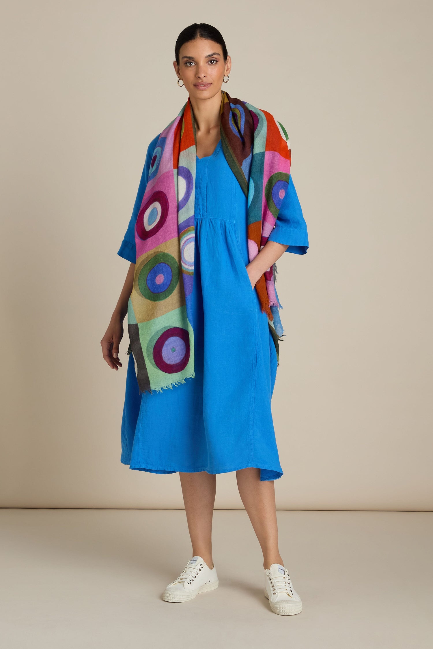 A person donning a bright blue dress, white sneakers, and the vivid Concentric Circles Wool Scarf featuring a geometric print stands against a plain background.