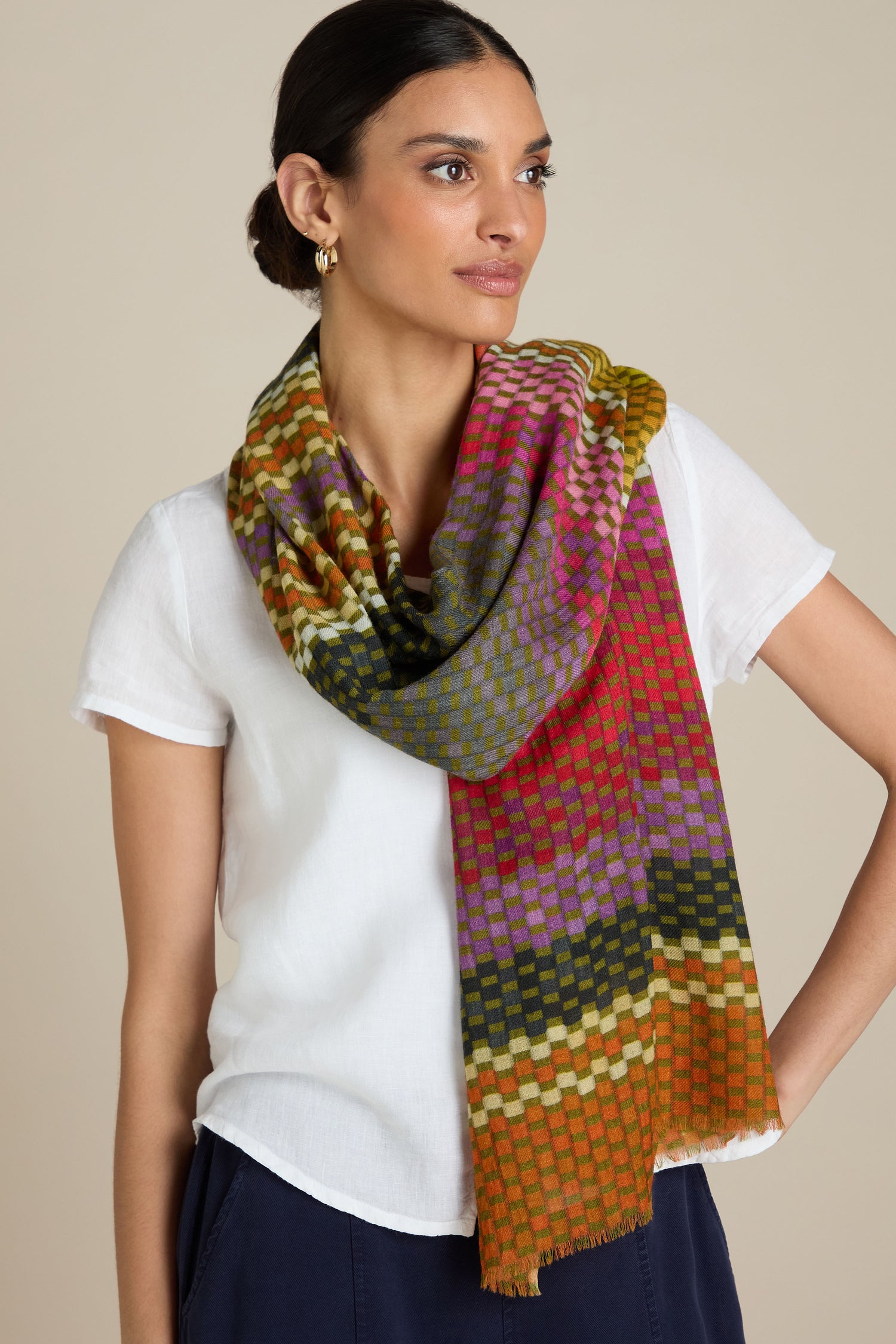 A person wears a white short-sleeve shirt paired with the Mini Squares Wool Scarf, which showcases vibrant gradient colors, elegantly draped around their neck. They are posing against a plain beige background.