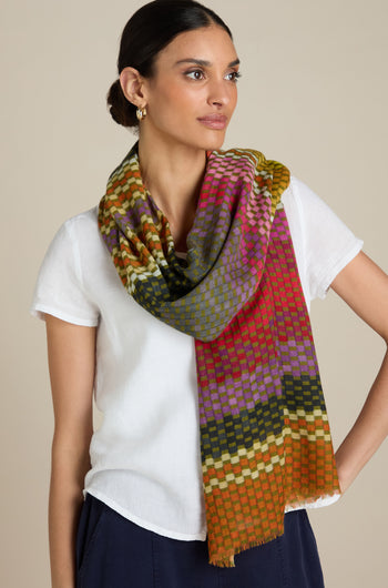 A person wears a white short-sleeve shirt paired with the Mini Squares Wool Scarf, which showcases vibrant gradient colors, elegantly draped around their neck. They are posing against a plain beige background.