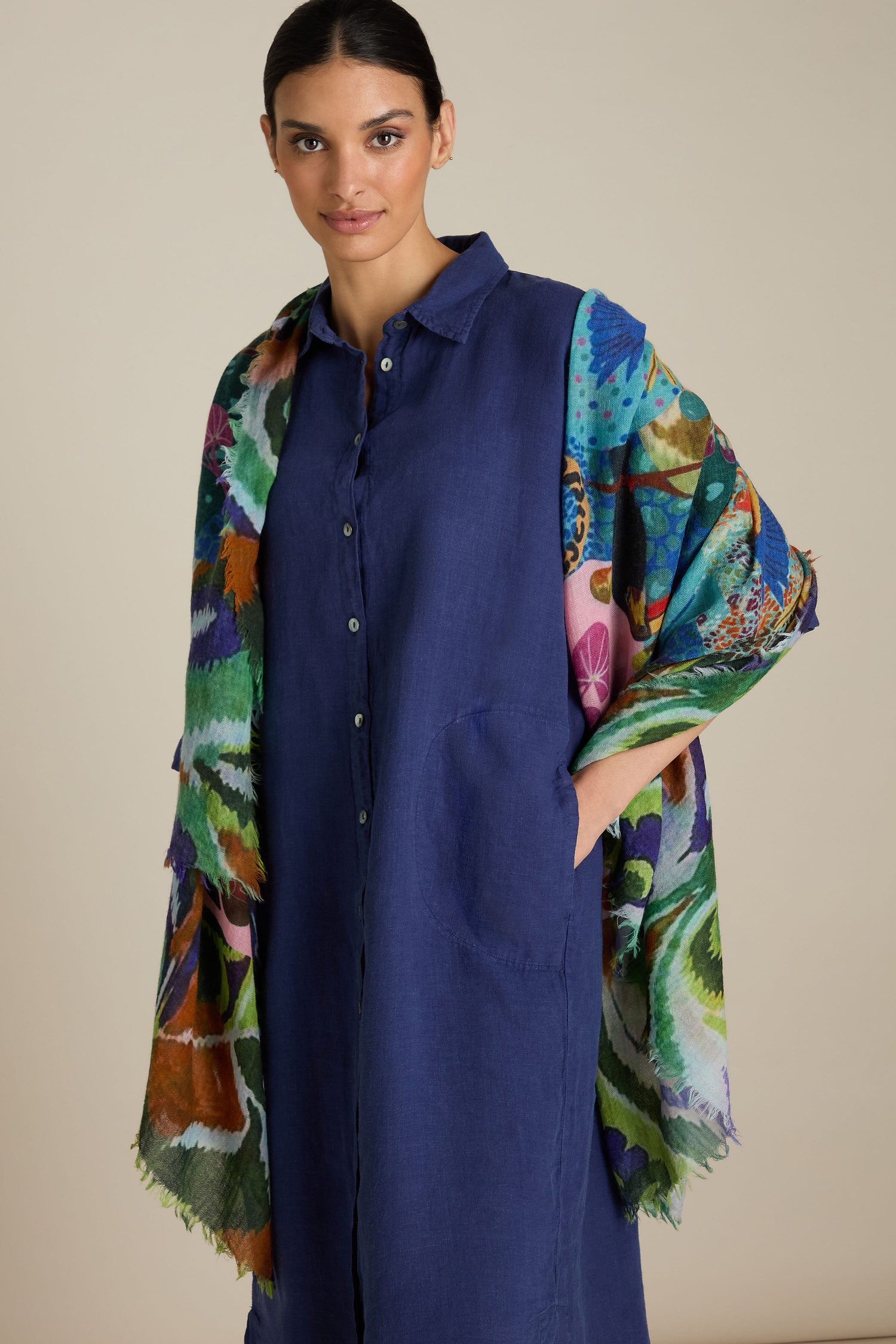 A woman stands wearing a long-sleeve blue dress with a button-down front and pockets, paired with the versatile Jungle Print Wool Scarf. She has dark hair pulled back and gazes directly at the camera.