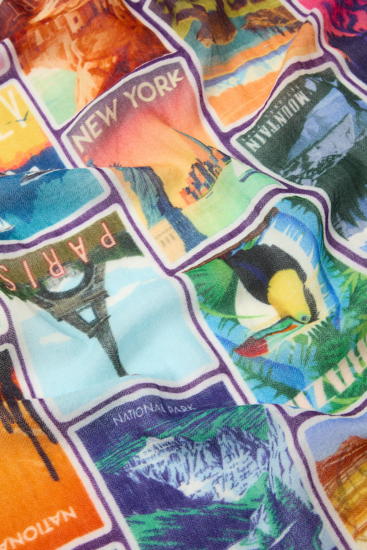 The Postcards From Around the World Wool Scarf showcases a multicolored pattern with illustrations of various famous landmarks, including New York, Paris, and National Parks, making it perfect for all-occasion styling and any wanderlust-inspired adventure.