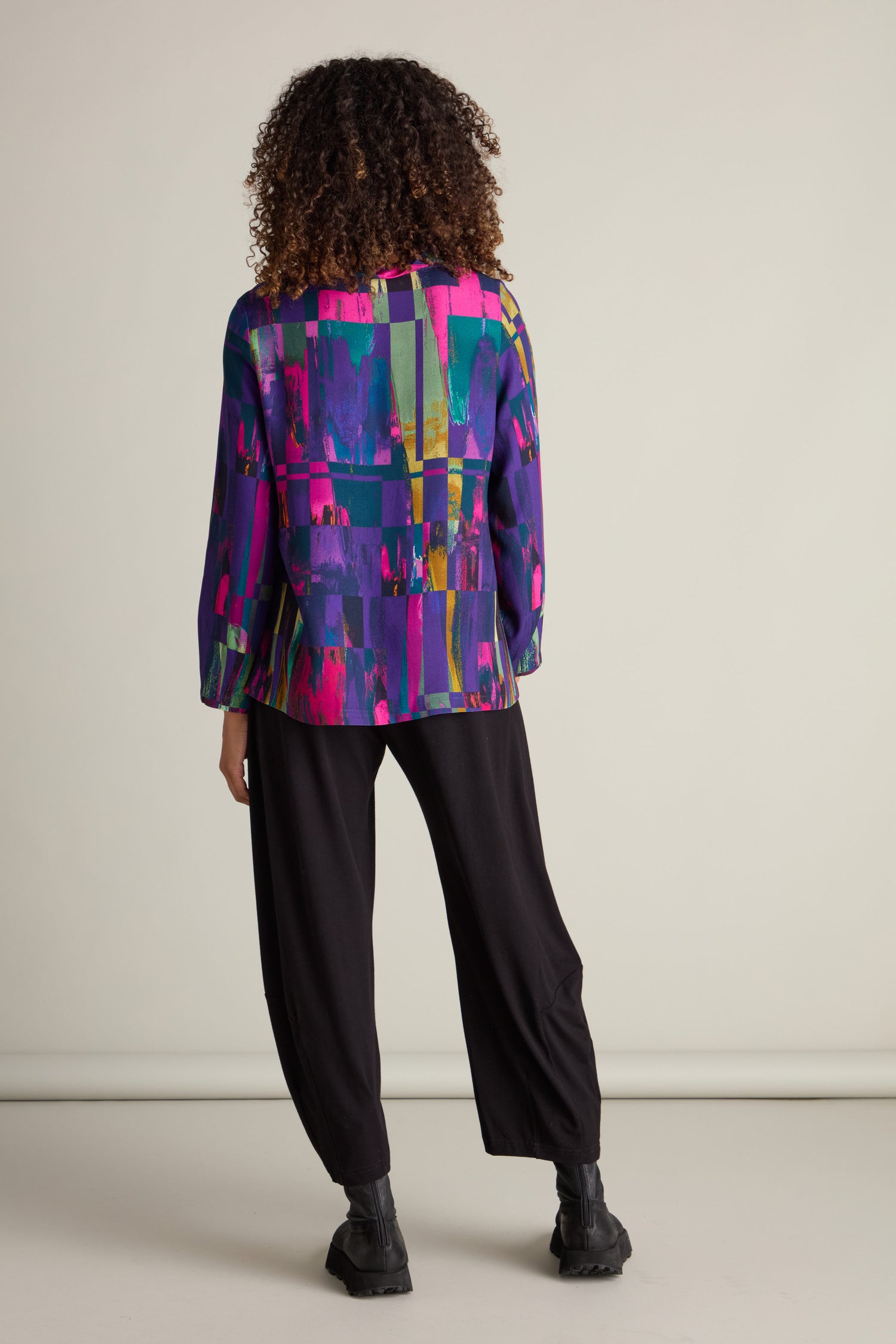 An individual with curly hair is seen donning an Aliza Block Print Top in vibrant jewel tones, paired elegantly with black pants and matching black boots, photographed from behind against a simple backdrop.