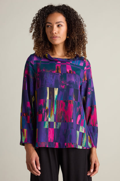 A woman with curly hair wears the Aliza Block Print Top, featuring a multicolored abstract pattern in jewel tones, paired with dark pants, standing against a plain background.