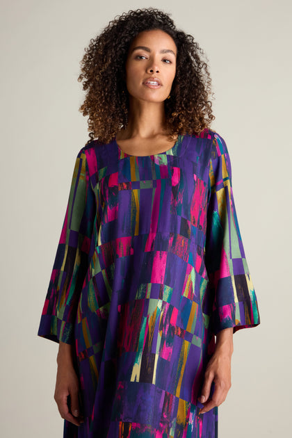 A woman with curly hair dons a Malina Block Print Dress, showcasing vibrant, abstract patterns and three-quarter length sleeves, as she stands against a plain background.