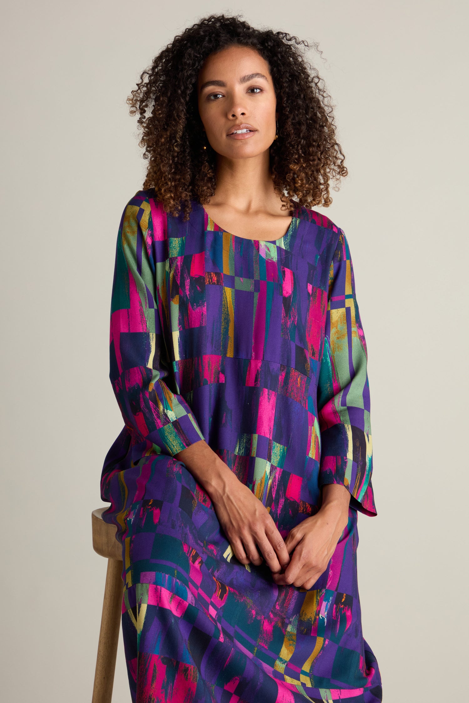 A person with curly hair wears a vibrant Malina Block Print Dress and sits on a wooden stool against a plain background.