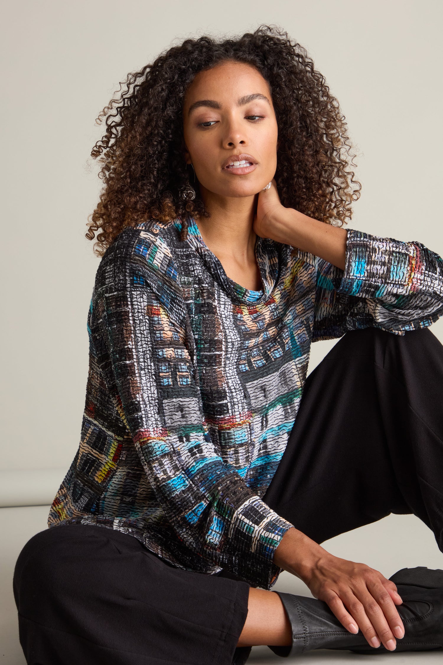 Clad in an Aliza Facade Print Jersey Top and black pants, a person sits with one hand on their neck and the other resting on their knee, introducing a hint of architectural intrigue against a plain backdrop.